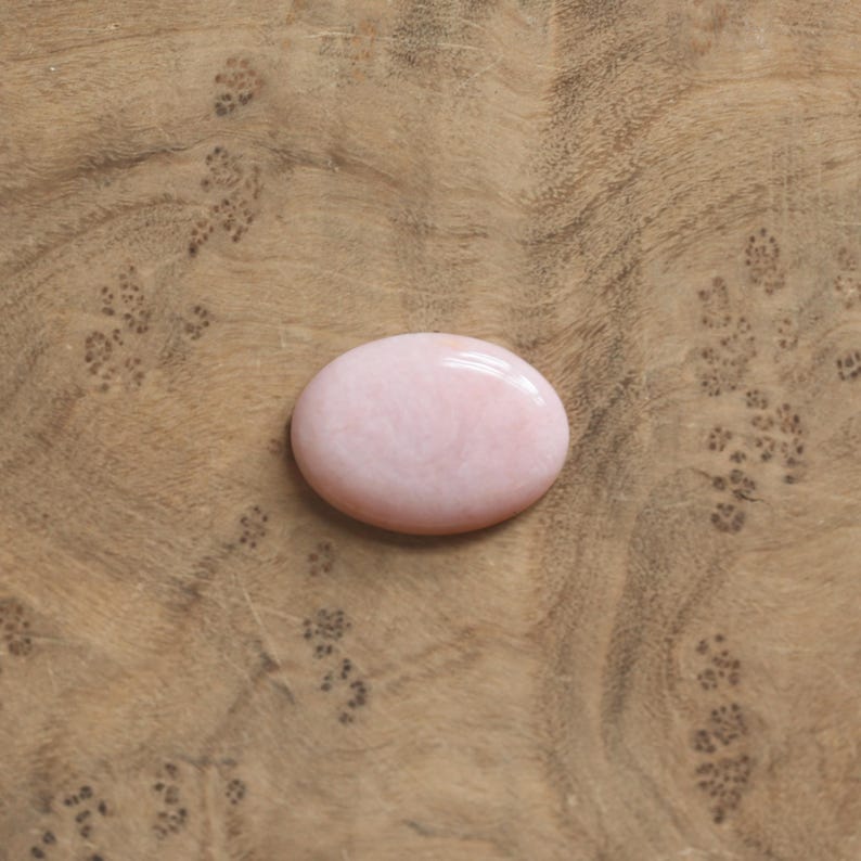 K&ouml;nnte beinhalten: Ein glatter, oval geformter rosa Edelstein mit polierter Oberfl&auml;che. Der Stein hat eine weiche, undurchsichtige hellrosa Farbe mit subtilen, wirbelnden Farbvariationen auf seiner abgerundeten Oberfl&auml;che, die das Licht gleichm&auml;&szlig;ig reflektiert.