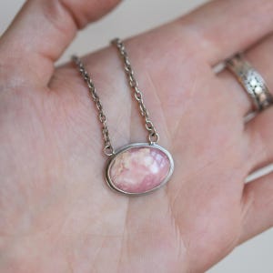 K&ouml;nnte beinhalten: Eine ovale Anh&auml;ngerkette mit einem glatten, polierten rosa-wei&szlig;en Stein, der in eine silberfarbene Metallfassung eingefasst ist. Der Anh&auml;nger ist an einer schlichten silberfarbenen Metallkette mit kleinen, gleichm&auml;&szlig;igen runden Gliedern befestigt.
