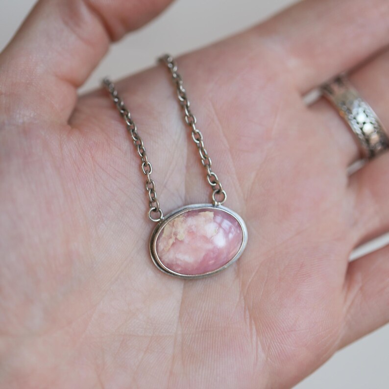 K&ouml;nnte beinhalten: Eine ovale Anh&auml;ngerkette mit einem glatten, polierten rosa-wei&szlig;en Stein, der in eine silberfarbene Metallfassung eingefasst ist. Der Anh&auml;nger ist an einer schlichten silberfarbenen Metallkette mit kleinen, gleichm&auml;&szlig;igen runden Gliedern befestigt.