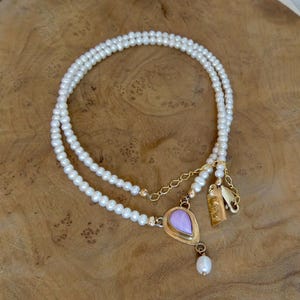 Puede incluir: Collar de perlas de doble hebra con un colgante dorado con una piedra lavanda en forma de l&aacute;grima. El collar tiene una cadena dorada y peque&ntilde;os dijes dorados. Las perlas son blancas.