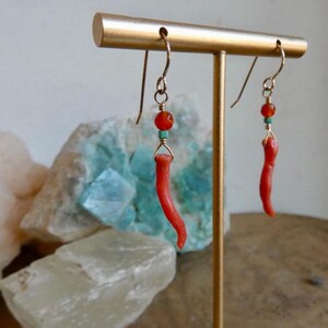 May include: A pair of dangle earrings featuring curved, horn-shaped red coral pendants suspended from gold-toned wire hooks. Each earring includes a small, round orange bead and a tiny turquoise-colored spacer bead above the coral element.