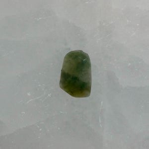 May include: A small, irregularly shaped gemstone with a polished surface, featuring a mottled pattern of deep forest green and translucent pale olive green hues. The stone has a smooth, rounded top edge and slightly faceted sides, displaying natural color variations throughout its semi-translucent body.