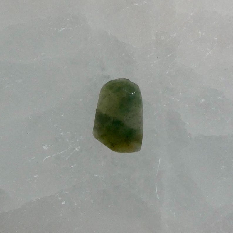 May include: A small, irregularly shaped gemstone with a polished surface, featuring a mottled pattern of deep forest green and translucent pale olive green hues. The stone has a smooth, rounded top edge and slightly faceted sides, displaying natural color variations throughout its semi-translucent body.