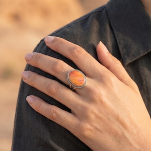 Könnte beinhalten: Ein Silberring mit einem großen, ovalen, orange-gelben Stein mit glatter, polierter Oberfläche. Der Stein ist in eine gedrehte Metallfassung im Seil-Design eingefasst, und die Ringschiene besteht aus zwei dünnen, parallelen Silberdrähten.