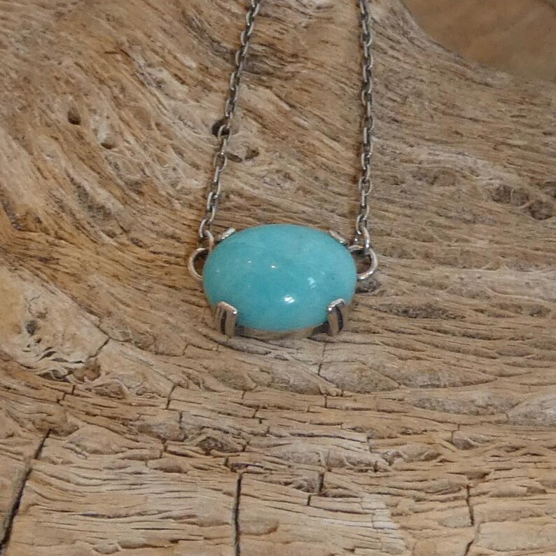 K&ouml;nnte beinhalten: Eine Halskette mit einem glatten, ovalen, hellblauen Edelstein in einer silberfarbenen Metallfassung mit Krappen. Der Anh&auml;nger ist an einer zarten silberfarbenen Metallkette mit kleinen, gleichm&auml;&szlig;igen ovalen Gliedern befestigt.