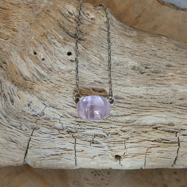 K&ouml;nnte beinhalten: Eine Halskette mit einem glatten, ovalen, durchscheinenden hellrosa Edelsteinanh&auml;nger, der an einer zarten, d&uuml;nnen Metallkette h&auml;ngt. Der Anh&auml;nger wird an beiden Seiten von kleinen Metallkrallen gehalten, die mit der Kette verbunden sind.