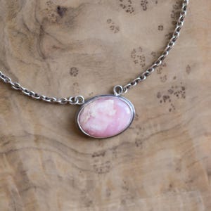 K&ouml;nnte beinhalten: Eine Halskette mit einem ovalen, rosa-wei&szlig;en Steinanh&auml;nger in einer silberfarbenen Metallfassung. Der Anh&auml;nger ist an einer silberfarbenen Metallkette mit kreisf&ouml;rmigen Gliedern befestigt, die an den Seiten der Steinfassung ansetzen.