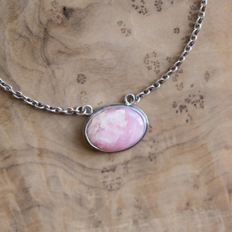 K&ouml;nnte beinhalten: Eine Halskette mit einem ovalen, rosa-wei&szlig;en Steinanh&auml;nger in einer silberfarbenen Metallfassung. Der Anh&auml;nger ist an einer silberfarbenen Metallkette mit kreisf&ouml;rmigen Gliedern befestigt, die an den Seiten der Steinfassung ansetzen.