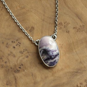 May include: A silver chain necklace with an oval pendant. The pendant features a polished stone with shades of lavender, white, and deep purple. The stone is set in a silver bezel.
