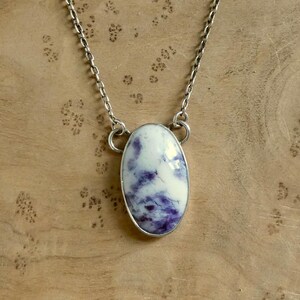 May include: A silver chain necklace featuring an oval pendant. The pendant has a white base with swirling purple patterns and a silver bezel. The necklace is displayed against a wooden background.
