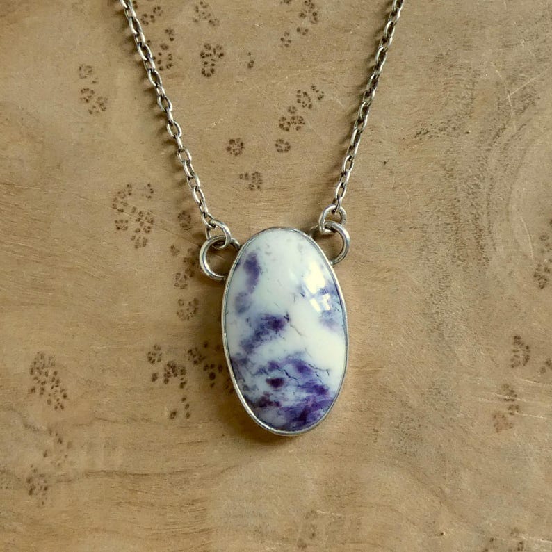 May include: A silver chain necklace featuring an oval pendant. The pendant has a white base with swirling purple patterns and a silver bezel. The necklace is displayed against a wooden background.