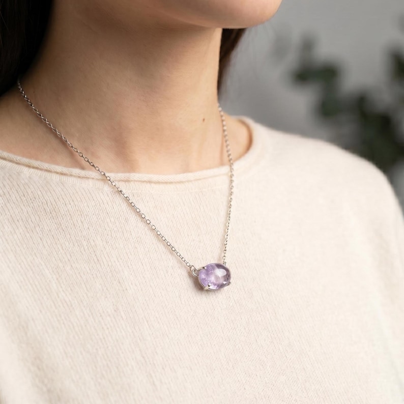 K&ouml;nnte beinhalten: Eine silberfarbene Halskette mit einer feinen Kette und einem einzelnen, ovalen, durchscheinenden violetten Edelsteinanh&auml;nger in einer schlichten Metallfassung. Der Anh&auml;nger sitzt mittig an der Kette, die mit einem Standardverschluss versehen ist.