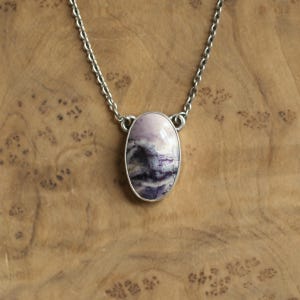 May include: A silver necklace featuring an oval pendant with a polished, marbled stone. The stone displays shades of lavender, purple, and white, set in a silver bezel. The chain is a delicate silver link. The necklace is displayed on a wooden surface.