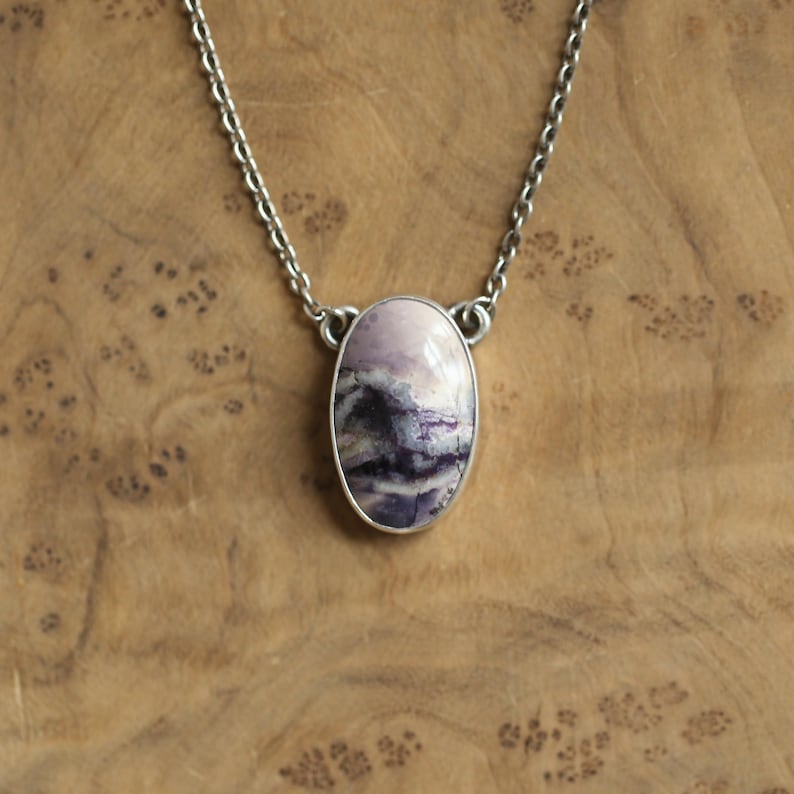 May include: A silver necklace featuring an oval pendant with a polished, marbled stone. The stone displays shades of lavender, purple, and white, set in a silver bezel. The chain is a delicate silver link. The necklace is displayed on a wooden surface.