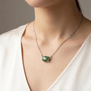 K&ouml;nnte beinhalten: Eine silberfarbene Halskette mit einem einzelnen, glatten, ovalen gr&uuml;nen Steinanh&auml;nger in einer Vier-Krappen-Fassung. Der Anh&auml;nger ist mittig an einer zarten, d&uuml;nnen Metallkette befestigt, die auf dem Schl&uuml;sselbeinbereich eines wei&szlig;en Kleidungsst&uuml;cks aufliegt.