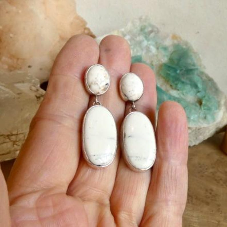 May include: A pair of drop earrings featuring two smooth, oval-shaped white stones with subtle grey veining. Each earring consists of a smaller circular stone at the top connected to a larger, elongated oval stone below, set in a simple metal frame.