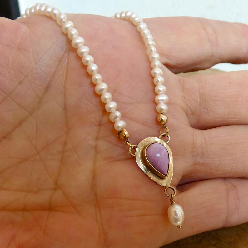 Puede incluir: Un delicado collar con una cadena de peque&ntilde;as perlas blancas. El colgante tiene forma de l&aacute;grima con una piedra de color p&uacute;rpura claro engastada en un metal dorado. Una sola perla cuelga de la parte inferior del colgante. El collar se muestra en una mano.