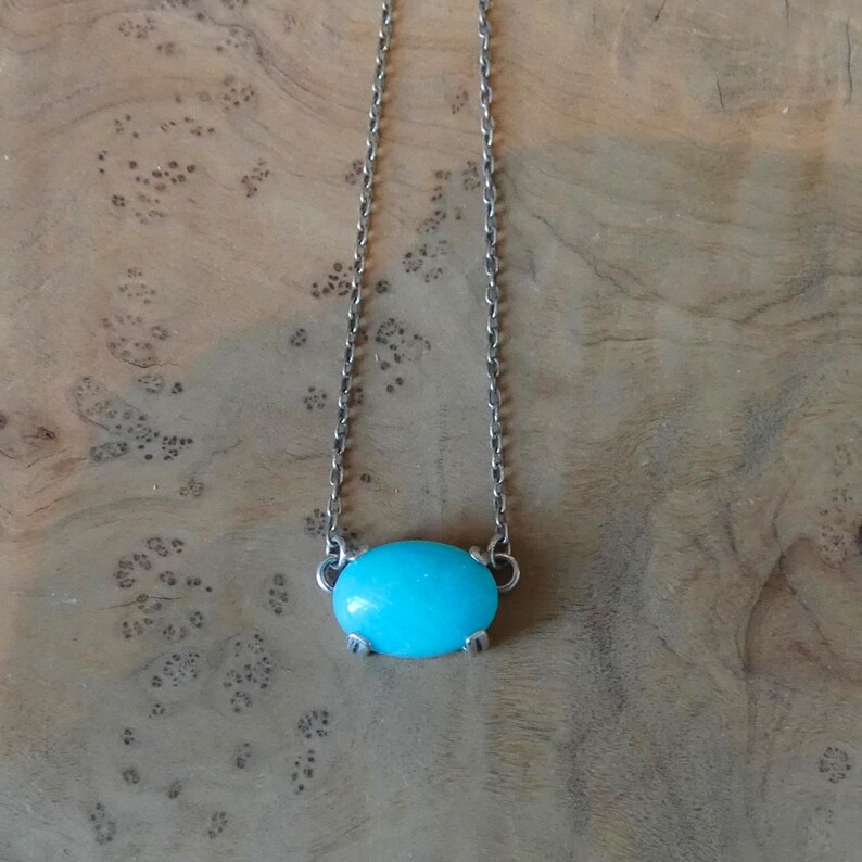 K&ouml;nnte beinhalten: Eine silberfarbene Metallkettenhalskette mit einem einzelnen, glatten, ovalen Anh&auml;nger in T&uuml;rkis. Der Stein wird von vier kleinen Metallkrallen gehalten, und die Kette ist direkt an den Seiten der Fassung des Anh&auml;ngers befestigt.