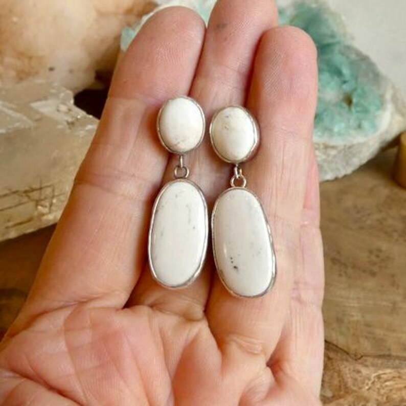 May include: A pair of drop earrings featuring two oval-shaped white stones with subtle grey inclusions, each set in a silver-toned metal frame. The design consists of a smaller oval stone at the top connected to a larger, elongated oval stone below.