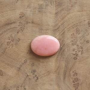 K&ouml;nnte beinhalten: Ein glatter, oval geformter rosa Edelstein mit polierter Oberfl&auml;che. Der Stein hat eine einheitliche, undurchsichtige hellrosa Farbe mit einem winzigen dunklen Punkt nahe der Mitte und zeigt einen nat&uuml;rlichen, abgerundeten Cabochon-Schliff.