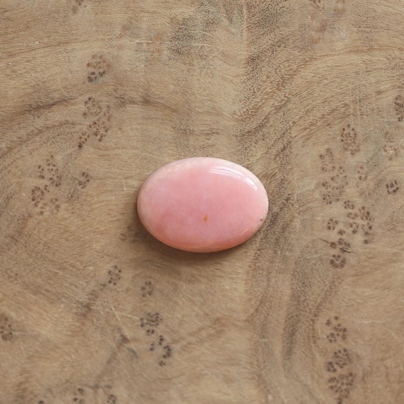 K&ouml;nnte beinhalten: Ein glatter, oval geformter rosa Edelstein mit polierter Oberfl&auml;che. Der Stein hat eine einheitliche, undurchsichtige hellrosa Farbe mit einem winzigen dunklen Punkt nahe der Mitte und zeigt einen nat&uuml;rlichen, abgerundeten Cabochon-Schliff.