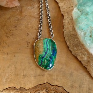 May include: A pendant necklace featuring a polished, irregular azurite-malachite gemstone with vibrant blue and green swirls, set in a simple silver-toned metal bezel. The pendant hangs from a thick, textured silver-toned chain link necklace.