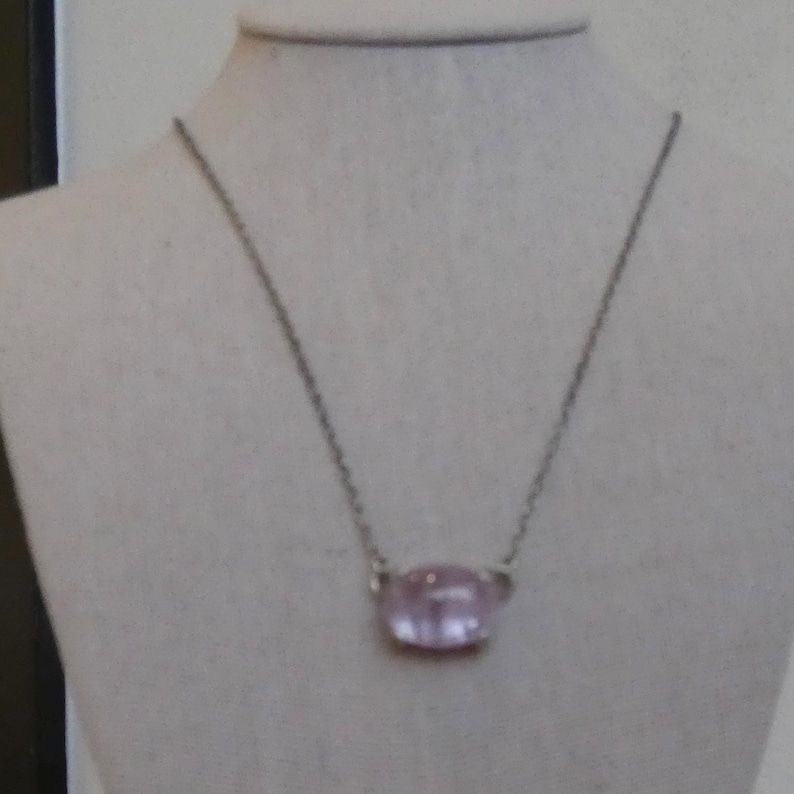 K&ouml;nnte beinhalten: Eine silberfarbene Metallkettenhalskette mit einem einzelnen, glatten, oval geformten hellrosa Edelsteinanh&auml;nger. Der Anh&auml;nger ist in einer einfachen Metallfassung befestigt, die direkt mit der feinen Gliederkette verbunden ist.