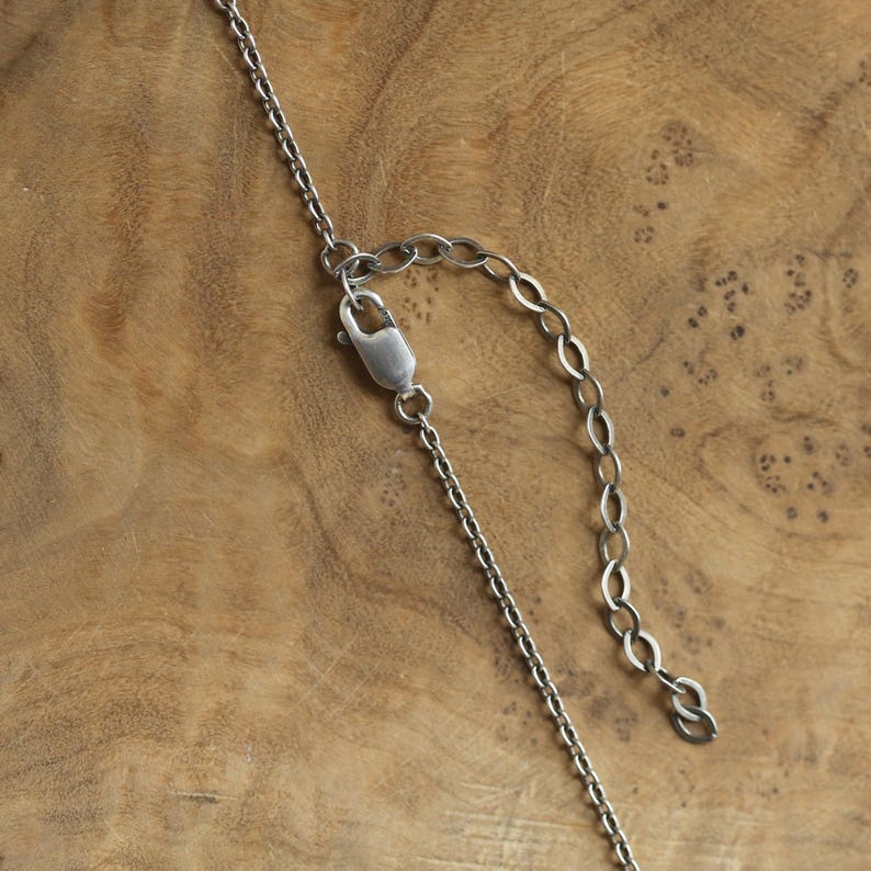 May include: Close-up of a silver-colored necklace chain with a lobster clasp and adjustable chain. The chain is made of small, interconnected links. The background is a wooden surface with a natural grain pattern.