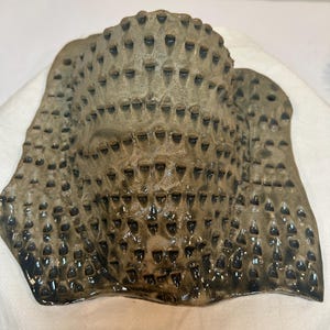 May include: A wall-mounted ceramic mask with a curved, protruding face shape. The surface is finished in a muted olive-green glaze and features a repeating pattern of small, dark, recessed triangular indentations across the entire exterior.