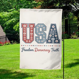 May include: A white garden flag with the word "USA" in red and blue letters, decorated with floral patterns. Below the letters are the words "Freedom Democracy Truth" and a row of stars.