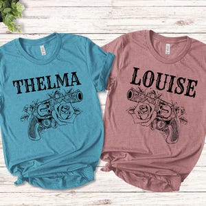 May include: Two t-shirts, one teal blue and one dusty rose, with a graphic of a gun and roses. The teal blue shirt says "THELMA" and the dusty rose shirt says "LOUISE".