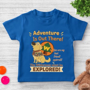 May include: A blue toddler t-shirt featuring a cartoon dog with the text "Adventure Is Out There!" and "The Wilderness must be EXPLORED!". The dog is wearing a brown collar and is set against a circular graphic.