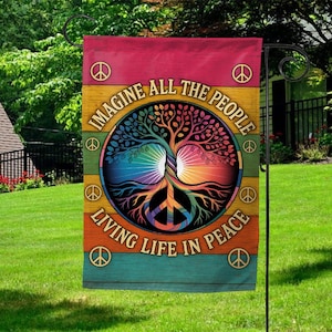 May include: A colorful garden flag with the text "IMAGINE ALL THE PEOPLE LIVING LIFE IN PEACE." The flag features a tree of life design with a peace symbol at the base. The background has horizontal stripes of red, yellow, orange, and teal.