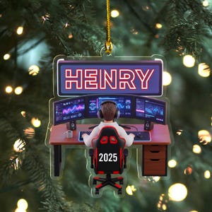 May include: A personalized Christmas ornament featuring the name "HENRY" in a neon-style sign. The ornament depicts a gamer at a desk with multiple monitors, a headset, and a gaming chair with "2025" on it. The background is a blurred Christmas tree.