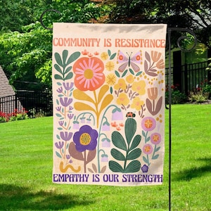 May include: A garden flag with a floral design and the text "Community is Resistance" and "Empathy is Our Strength". The flag is cream colored with a variety of flowers in shades of pink, purple, yellow, and green. There is a butterfly and a ladybug on the flag.