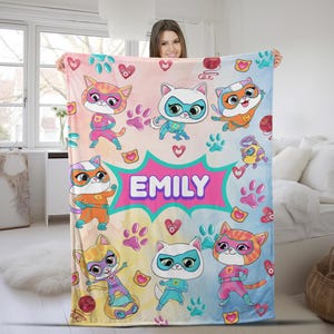 May include: A pink, blue, and yellow fleece blanket with a colorful pattern of paw prints, hearts, and superhero cats. The blanket features the name "EMILY" in a pink speech bubble.