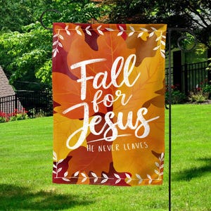 May include: A decorative garden flag with a fall theme. The flag features a background of orange and yellow autumn leaves. White script reads "Fall for Jesus" with "He Never Leaves" below. The flag is hanging on a black metal stand.