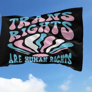 May include: A black flag with the text "TRANS RIGHTS ARE HUMAN RIGHTS" in a colorful, wavy font. The text is surrounded by a stylized, wavy design in pink, blue, and white.