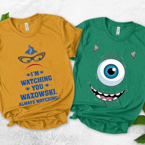 May include: Two short-sleeved t-shirts. One is mustard yellow with a graphic of a character's face, glasses, and the text "I'M WATCHING YOU WAZOWSKI, ALWAYS WATCHING...". The other is green with a cartoon monster face.