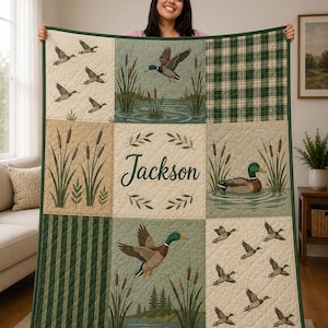 May include: A quilted blanket featuring a grid of nine panels with illustrations of mallard ducks in flight and on water, cattails, and green plaid patterns. The center panel displays the name Jackson in dark green script surrounded by a leaf wreath.