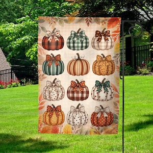 May include: A decorative garden flag featuring a pattern of twelve pumpkins with various designs and bows. The pumpkins are in shades of orange, brown, green, and white, with plaid, floral, and polka dot patterns. The flag is displayed outdoors on a black metal stand.
