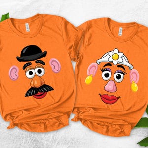May include: Two orange t-shirts with cartoon Mr. and Mrs. Potato Head faces. One shirt features Mr. Potato Head with a black hat and mustache. The other shirt features Mrs. Potato Head with a white hat and earrings.
