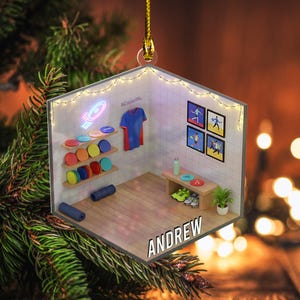 May include: A miniature disc golf scene ornament. The interior features shelves with colorful discs, a blue shirt, framed artwork, and a bench with shoes. The name "ANDREW" is displayed on the floor. The ornament is suspended by a gold string.