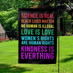 May include: A black garden flag with rainbow text that reads "Science is real, Black Lives Matter, No Human is Illegal, Love is Love, Women's Rights are Human Rights, Kindness is Everything."