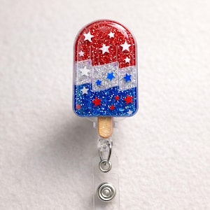 May include: A red, white, and blue popsicle-shaped badge reel with glitter and star accents. The badge reel has a wooden stick and a clear plastic retractable cord with a metal clip.