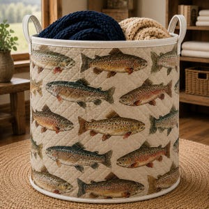 May include: A cylindrical quilted storage basket featuring a repeating pattern of brown and speckled trout fish on a light beige background. The basket has two white fabric handles on the sides and contains a dark blue and a tan folded blanket.