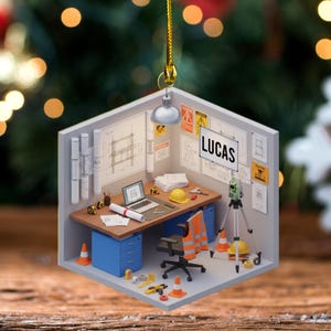 May include: A detailed ornament depicting an architect's office, complete with a desk, laptop, blueprints, and safety equipment. The name "LUCAS" is displayed on a whiteboard. The ornament is a miniature diorama of a workspace.