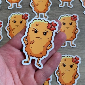 May include: A collection of 12 cartoon stickers featuring a yellow, angry, anthropomorphic chicken nugget with a red flower on its head. The stickers are on a wood surface.