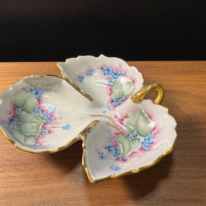 May include: A leaf-shaped, three-sectioned porcelain dish with a gold handle and trim. The dish is painted with green leaves, blue flowers, and pink accents. The dish is sitting on a wooden surface.