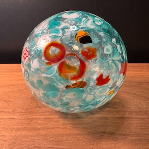 May include: A decorative glass sphere with a turquoise base color and white, orange, and red accents. The sphere has a round shape and sits on a wooden surface. The glass has a textured appearance.